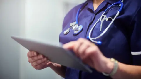A close up image of a medical professional typing on a tablet with a stethoscope around her neck. You cannot see her face.