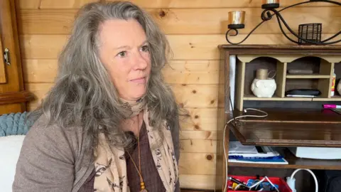 A woman in a beige cardigan with long grey hair sorts through a box of medication at a table. She is looking at a small blister pack of tablets which she is holding in her right hand.