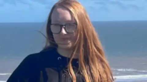 Cassidy standing outdoors on a sunny day with the sea and a sandy shoreline visible in the background. She is wearing glasses, a black zip‑up jacket and matching black trousers. Her long, ginger hair falls over one shoulder.
