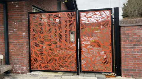 BBC The tall gate at the entrance to the Lilias Centre, which is orange with a leaf pattern. It sits between a red brick building and a red wall.