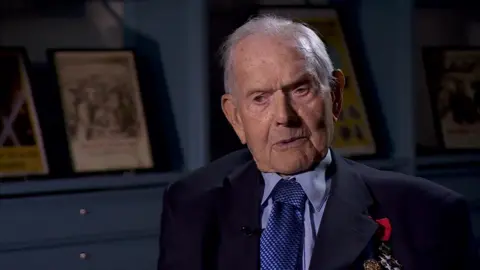 BBC Percy Chafer, an elderly man in a light blue shirt and tie and navy blue jacket featuring military medals. He sits in a blue room at the National Archives with framed historical posters behind him.