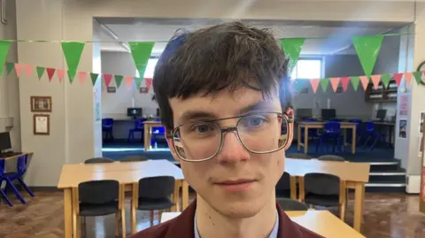 Patrick, a teenage boy with dark hair, large rimmed glasses, who is wearing a claret blazer and a light blue shirt. There are brown tables and green bunting in the background.