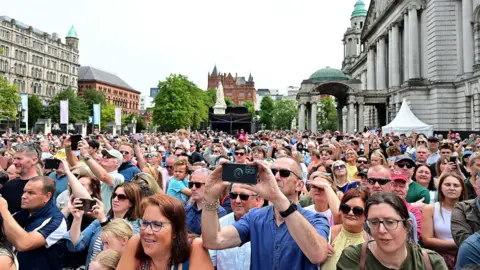 Pacemaker A large crowd of people stand in the grounds of Belfast City Hall, which is surrounded by tall buildings. Many are wearing sunglasses and are using mobile phones to record what they see on a stage in front of them.