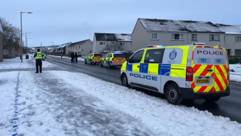 A police van and an officer in a hi-vis vest over black clothing standing next to a snow path in Inverness.