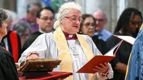 The Right Reverend Sarah Clark has white hair in a ponytail. She is wearing red and clear framed glasses and is taking her oath, reading from a red folder, which she is holding. She is wearing clerical clothing. 