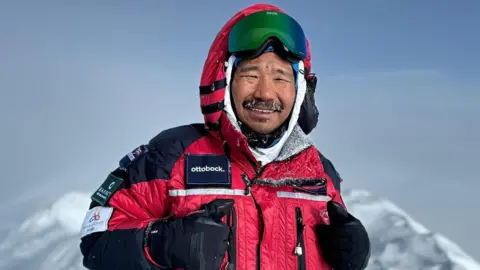 Hari Budha Magar Hari Budha Magar MBE, wearing a red and black coat and smiling at the camera. He is stood atop a mountain with a grey sky in the background.