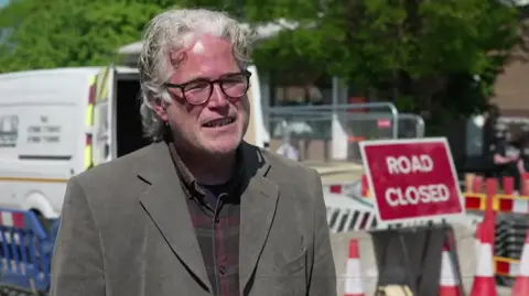 A grey haired man with glasses stands in front of a 'road closed' sign at the Loose Road closure site.
