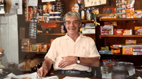 Dave Masters with grey hair and a pale cream shirt is sitting at the counter in his village shop, smiling at the camera. There are sweets and other food items stacked on shelves behind him. The photo was taken a few decades ago, with old branding visible on Quality Street boxes, Fruit Pastel packets and Fray Bentos pies, among other items.
