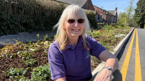 Patti Pinto, a woman wearing a purple polo shirt, is sitting on the curb at the side of a road with double yellow lines along it. Next to her is a planted flowerbed. There are trees and buildings in the distance behind her.