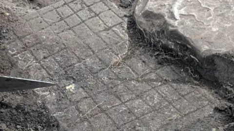 The Roman board game when it was still in the ground. It is a grey slab of rock with a grid pattern. Someone is holding a knife to the left of the photo and a large grey rock to the right.