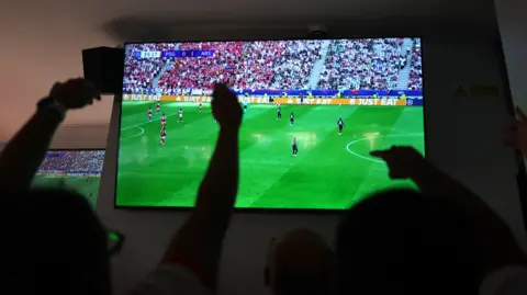 Getty Images A football match shown on a large TV. Viewers are seen from behind cheering, with their arms in the air, their figures are silhouettes. 