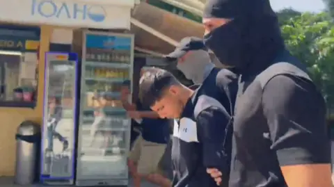 Sharif Ahmadzai leaving the court between two men dressed in black with face coverings, who are holding his arms.