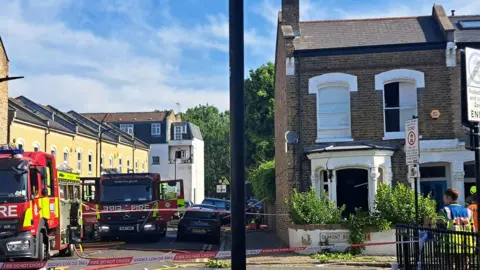 A house with a blown out window on a residential street, with fire engines parked along the road 