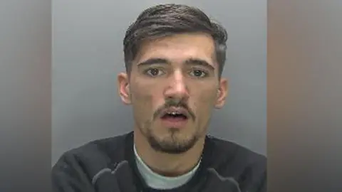 A man with his mouth slightly open in a police head-and-shoulders custody photo. He is wearing a grey sweatshirt and a white top underneath.