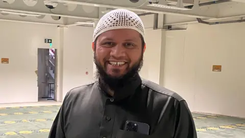 Abdul Basith is a Muslim man wearing a brown tunic and a white head covering with a beard. He is standing in a large prayer hall and smiling. He has a phone in his pocket 