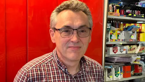 GUY CAMPBELL/BBC Shop keeper wearing glasses standing behind his counter