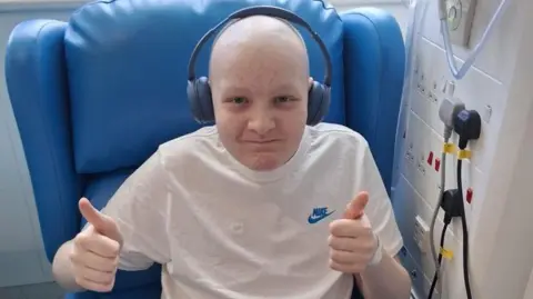 A boy in a hospital chair. He is bald, wearing noise-cancelling headphones, and gives two thumbs-ups to the camera.