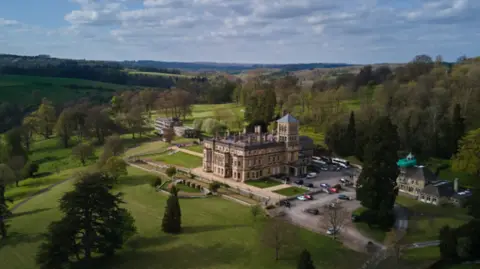 Drone photograph of a mansion, which serves as a private school, surrounded by parkland and woodland in the Cotswolds countryside. There is a car park to the side and manicured lawns surrounding the building.