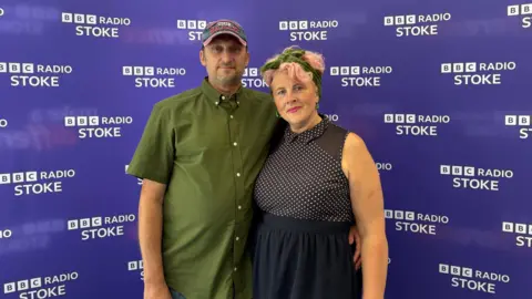 A man with a blue denim hat on is smiling and wearing a khaki green shirt. He has his arm around his wife, who has pink hair with a khaki green headband and is wearing a black and white dress with a spotty bodice and plain black skirt. They are standing in front of a purple backdrop that says "bbc radio stoke"