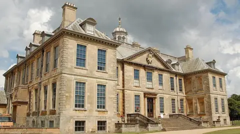 Getty Images A large historic stone mansion with tall sash windows, ornate architectural detailing and multiple chimneys, set against a dramatic cloudy sky. The building features a central entrance with steps leading up to it, flanked by symmetrical wings. A gravel path curves across the neatly kept lawn in the foreground.