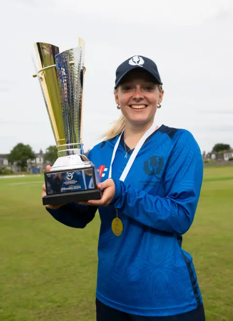 Cricket Scotland Niamh Muir, with blonde hair, is in a Scotland top and wearing a blue cap with a thistle on it. She is holding up a large trophy while wearing a medal around her neck