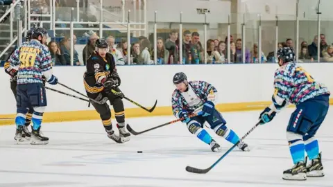 Five ice-hockey players on the ice holding hockey sticks and chasing a puck, with a crowd looking through perspex glass in the background