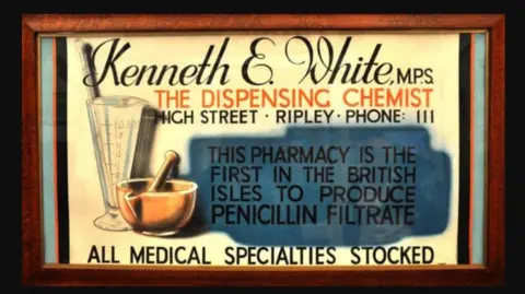 An old fashioned sign reading Kenneth E White MP, the dispensing chemist, high street, Riple, Phone III. This pharmacy is the first in the British Isles to produce penicillin filtrate. All medical specialities stocked.