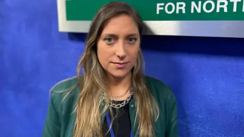 Fernanda Hermosilla standing in front of an Equality Commission for Northern Ireland sign.  She has long, straight, dark-fair hair and is wearing a green jacket over black top and two silver necklaces. 