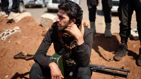 A young man with a light machine gun resting on his knees sits on a pile of earth and rubble. He's looking away to the side, absent-mindedly.