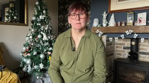 Pauline Stonehouse looking into the camera. She has red hair and is wearing red glasses and a green shirt. She's sitting in a living room with a Christmas tree behind her.