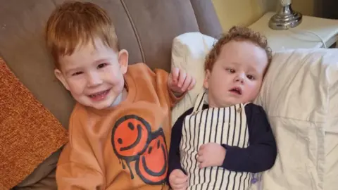 Bobby, left, is smiling directly at the camera. He has ginger hair and is wearing an orange jumper with smiley faces on it. Harry is laying on a white pillow, he has curly ginger hair. He is wearing white stripy dungarees and a navy top underneath. 