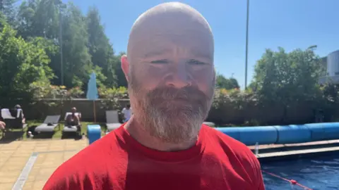 Jim McParland with a beard is standing up by the pool. He is wearing a red top and looking at the camera