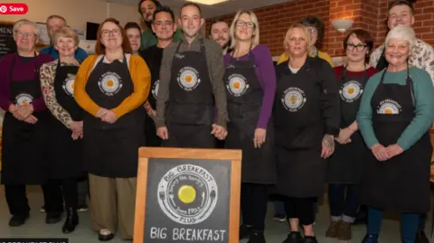 Big Breakfast Plus Recipients gathered together wearing black aprons with the Big Breakfast Plus logo on the front, all smiling to camera