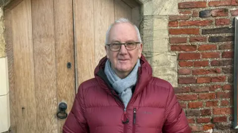 BBC/Tim Dale Mike Calvert has short grey hair and is wearing a dark burgundy coat and grey scarf. He has black framed glasses on and is standing outside a door to the Red Tower in York.