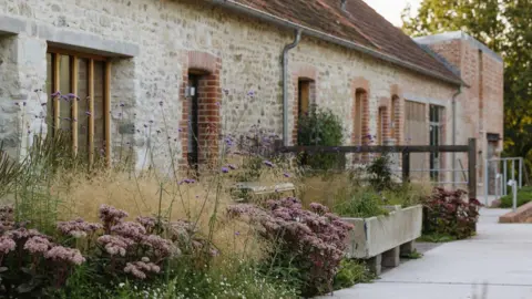 Eva Nemeth Un chemin longe un long bâtiment en pierre d'un étage avec un toit en pente. Un parterre de fleurs longeant le chemin regorge de graminées ornementales et de fleurs sur un thème rose et violet.