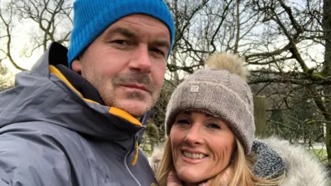Ross Kaye A selfie of Ross and his wife Tracey. Ross is wearing a hooded raincoat and a blue beanie. Tracey is wearing a pink rollneck sweater, a coat with a fluffy hood and a beige bobble hat.
