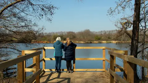 The Greensand Trust Two women stand on the new wooden boardwalk overlooking the lakes. It is a sunny day.