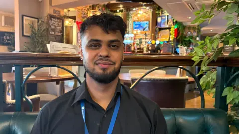 Martin Heath/BBC Nikhil Shukla wearing a black shirt and blue lanyard sits on a green leather bench inside a pub or restaurant. Wooden tables, upholstered chairs and a well-stocked bar with illuminated beer taps are visible in the background. Decorative plants frame the scene, and warm lighting creates a cosy atmosphere. The image is a centred portrait taken indoors with the background softly out of focus.