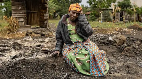 AFP A woman in an orange head scarf looks at the camera holding her head with one hand.