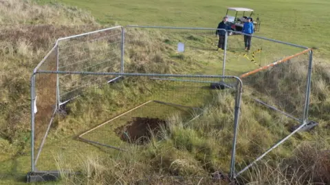 BBC A hole in the ground, from above, surrounded by six metal fences. The hole is in the middle of a grassy area which is surrounded by longer rough grass like the type found in sand dunes. A fence panel is resting on top of the hole that is in the middle of the image.