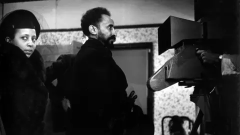 Haile Selassie and his daughter Princess Tegnagnework walking across a room being filmed by a large camera