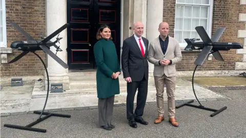 John Healey standing in between a male and female Helsing representatives and models of their Strike Drone HX-2.