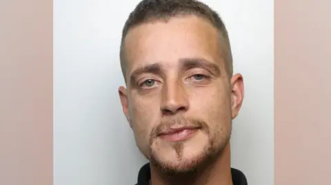 Declan Marsden has short brown hair and a moustache and short beard. He is looking at the camera and is wearing a black top.