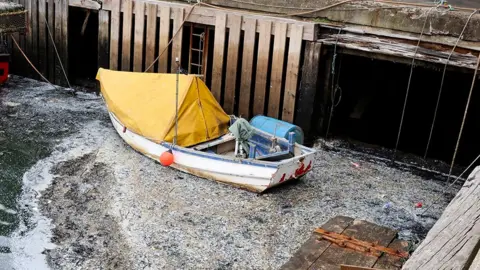 Leah Pattem Corner of a quayside with a moored small boat with yellow tarpaulin cover. There are wooden panels leading down from the quayside to the water, along with assorted ropes and a metal ladder. In the water, in the corner, a slick of nurdles have collected and, from a distance, look like frothy scum on the water.