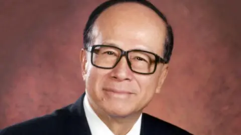 Li Ka Shing Foundation Sir Ka Shing Li smiling at the camera against a red background. He is wearing black square framed glasses, a black blazer, white shirt and has black hair.