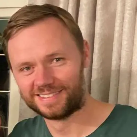 Ministry of Defence/PA Wire Lt Cdr Chris Gayson. He his pictured wearing a green top and is looking in the direction of the camera. He is smiling. He is standing in front of a curtain. 