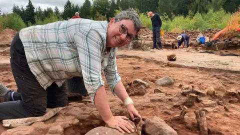 Catherine Wood in a checked shirt, leaning over some rocks