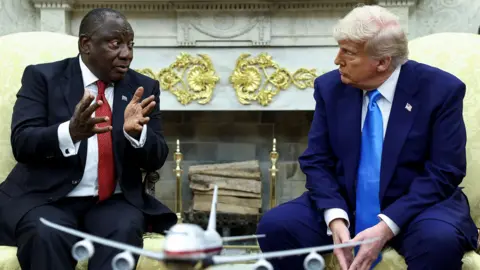Ramaphosa gestures as he responds to a question from Donald Trump in the Oval Office on 21 May. They both wear suits and tie and are sitting down on white armchairs.
