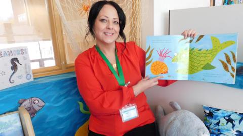 Adult learner Joanne Hassan, who has dark hair and is wearing a red blouse, standing smiling holding a brightly coloured children's book with fish and snail illustrations. She is surrounded by soft toys and paintings of a fish and seahorse