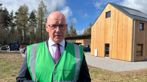 John Swinney wearing a suit, tie and hi-viz green jacket standing in front of a wooden house and trees.